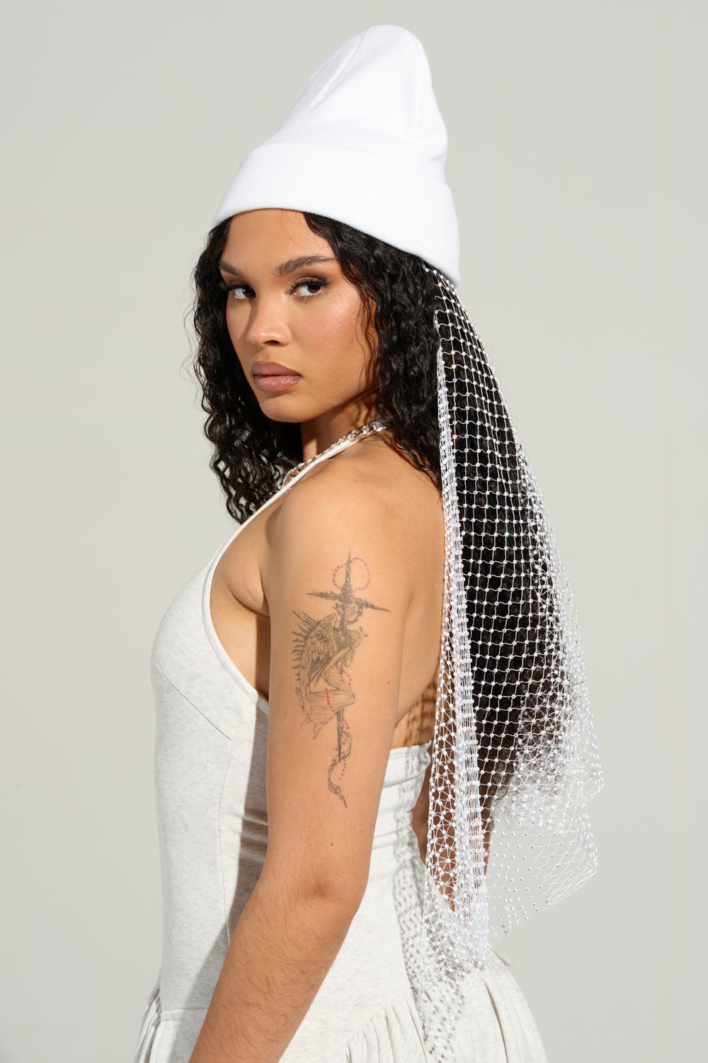 A person with long, curly hair is wearing the TRAIL OF RHINESTONES BEANIE, featuring rhinestones and a mesh veil at the back. They are dressed in a light-colored sleeveless top that reveals a detailed tattoo on their upper arm against a plain background. 2