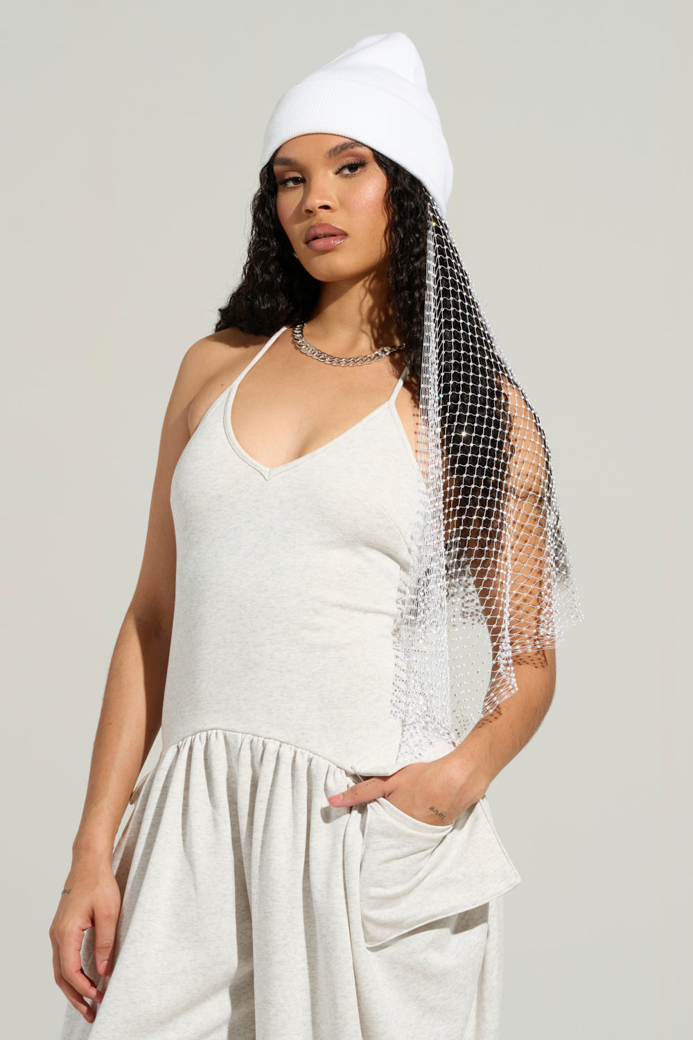 A person with long curly hair wears the TRAIL OF RHINESTONES BEANIE, a sleeveless light gray dress, and a silver chain necklace. They have a serious expression and a sparkling netting accessory draped over their shoulder. 3