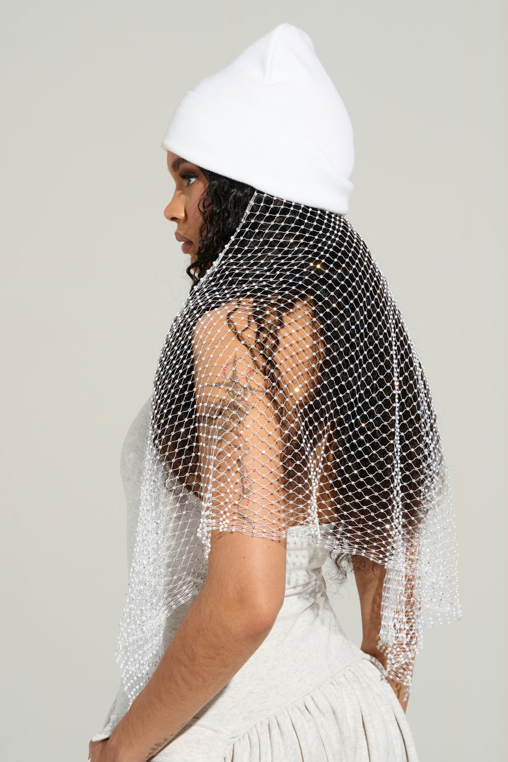 A person with long, curly hair is looking to the side while wearing a TRAIL OF RHINESTONES BEANIE and a mesh shawl over a light-colored outfit, set against a plain gray background. 4