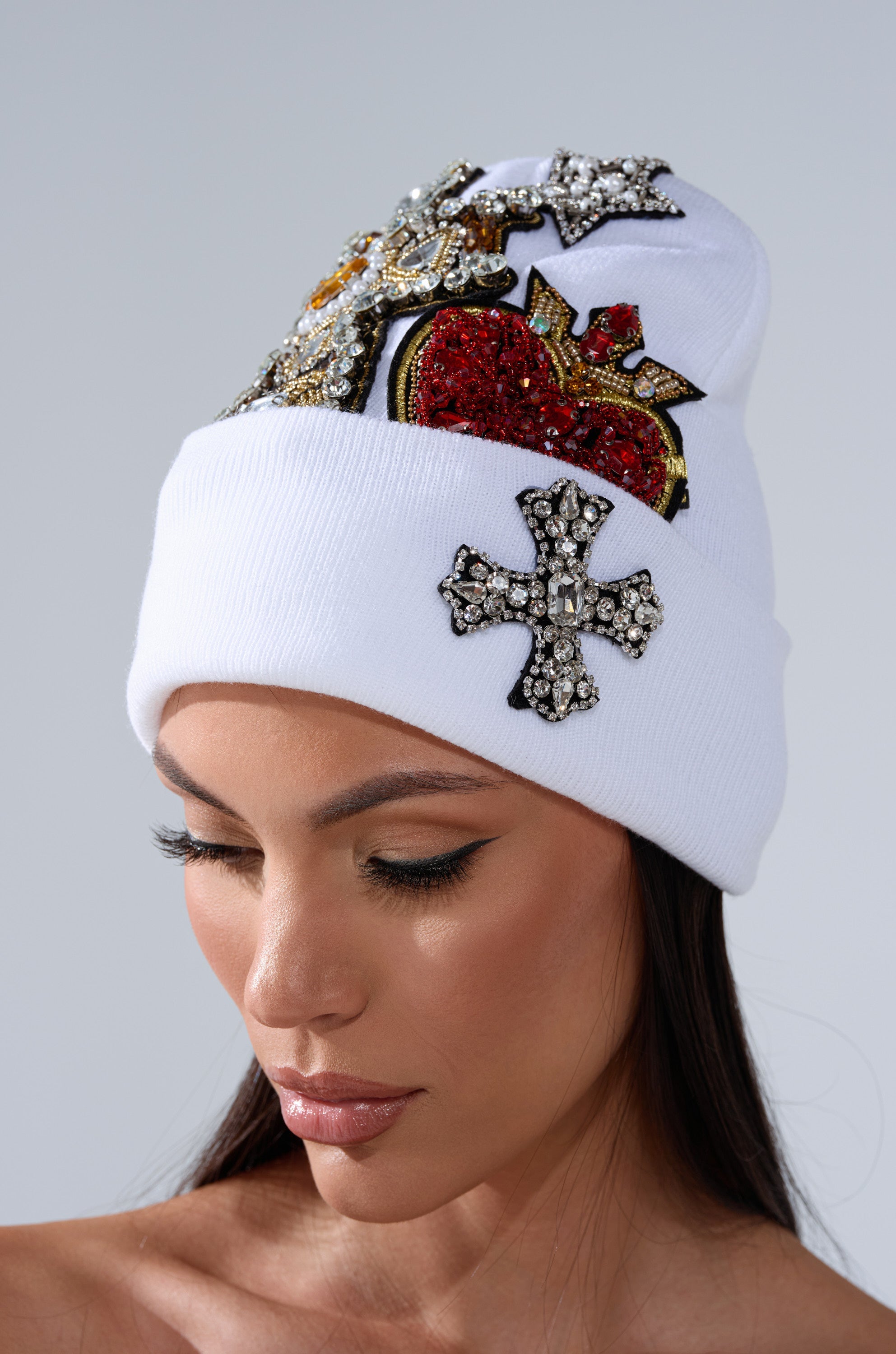 A woman wears the NO ONE ELSE LIKE ME BEANIE, a white rhinestone hat decorated with jeweled patches—a red heart, stars, and a cross—while looking downward against a plain light gray background. 0