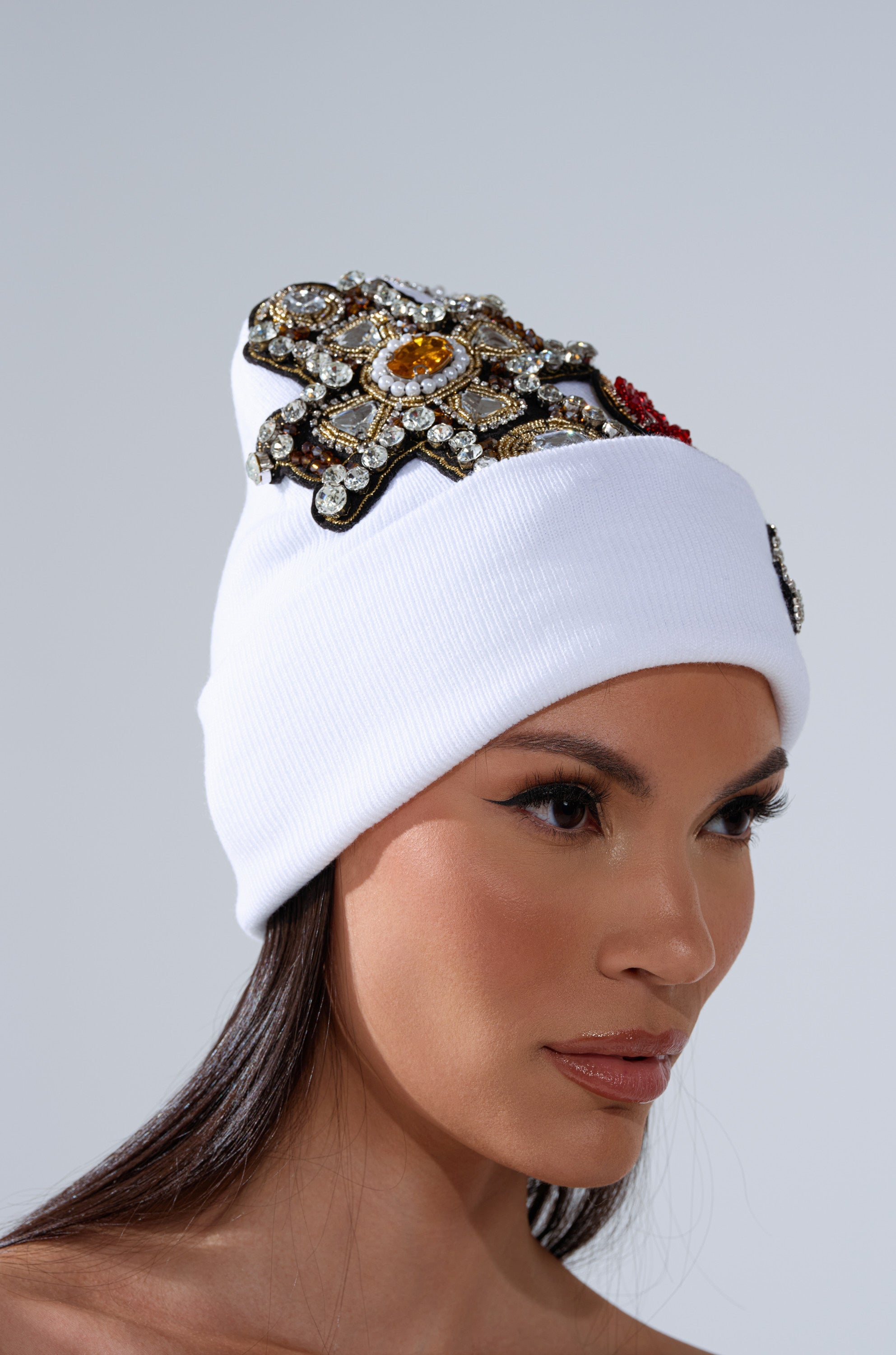 A woman with long dark hair and subtle makeup wears the NO ONE ELSE LIKE ME BEANIE, a white beanie featuring a large ornate rhinestone embellishment, as she faces slightly to the left. 2