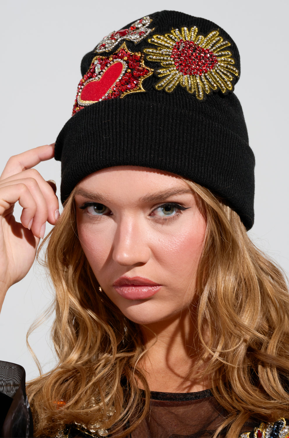 A person with long, wavy hair wears the REGENCY PATCH BEANIE, a black beanie decorated with colorful rhinestone-embellished heart and flower patches. They gently hold the edge of the beanie while gazing slightly to the side against a plain background.