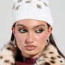 A person sporting the ASAP Vibes Embellished Beanie from the AKIRA Label and a leopard print faux fur coat radiates iconic vibes. They accessorize with hoop earrings and enhance their look with bold makeup—eyeliner and blush—as they pose against a plain background.