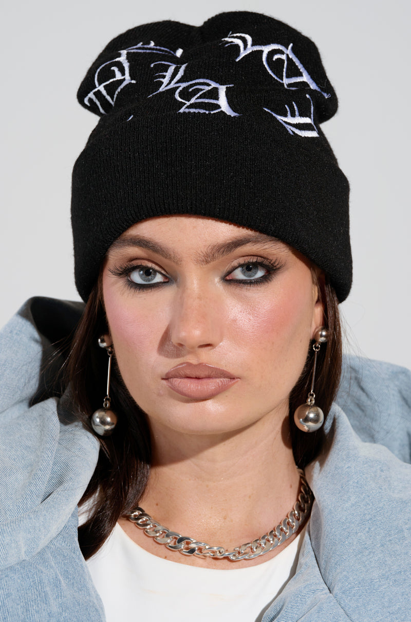 A person with long dark hair is wearing a CITY SHII embroidered beanie, metallic pearl earrings, and a silver chain necklace. Their dramatic eye makeup and neutral lip color complement the denim jacket draped over their shoulders, perfectly capturing the City Shii aesthetic.