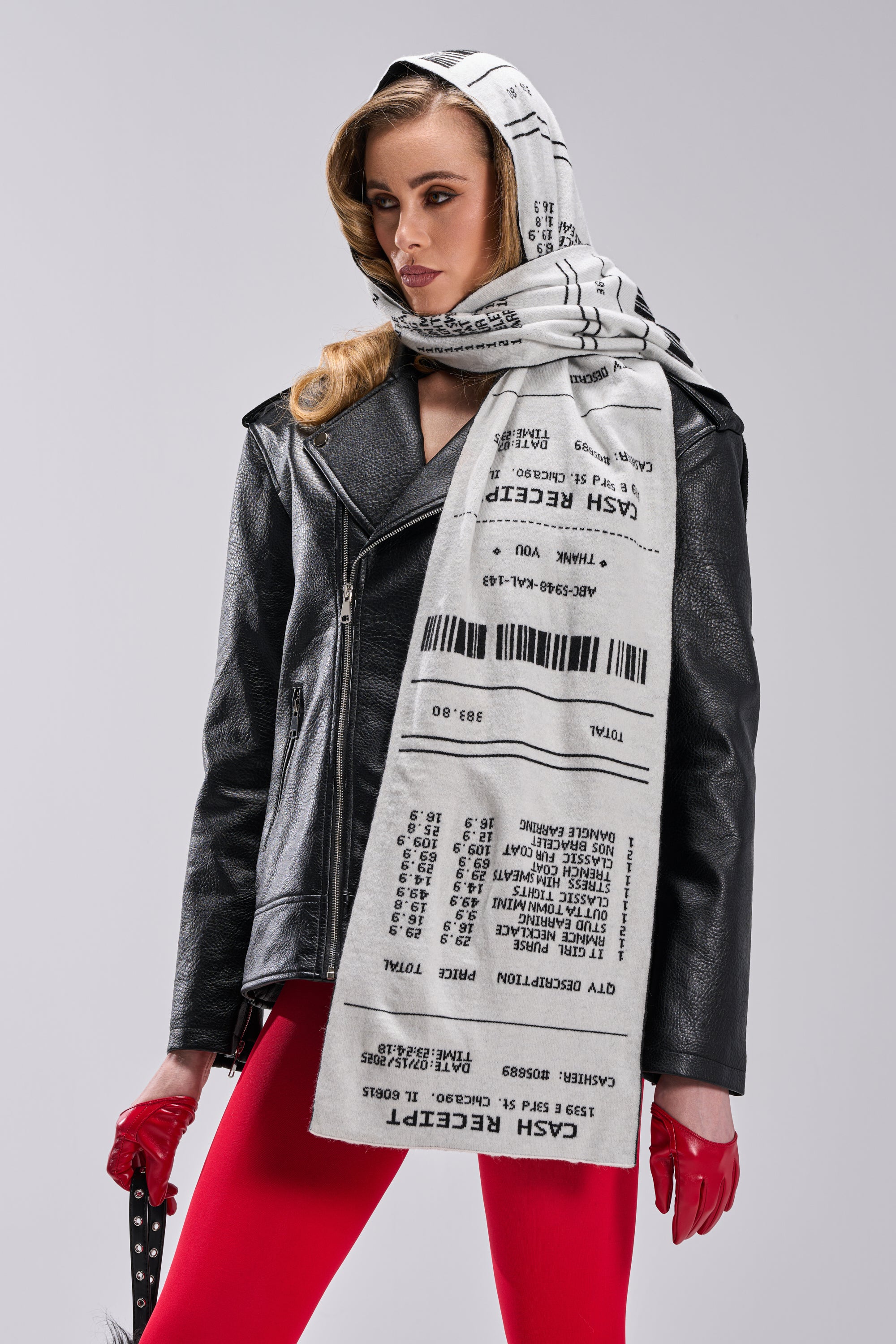 A woman with blonde hair and red gloves models the CHECK THE RECEIPT SCARF, a large accessory featuring a cash register receipt print, styled with a black leather jacket and red pants against a plain background.