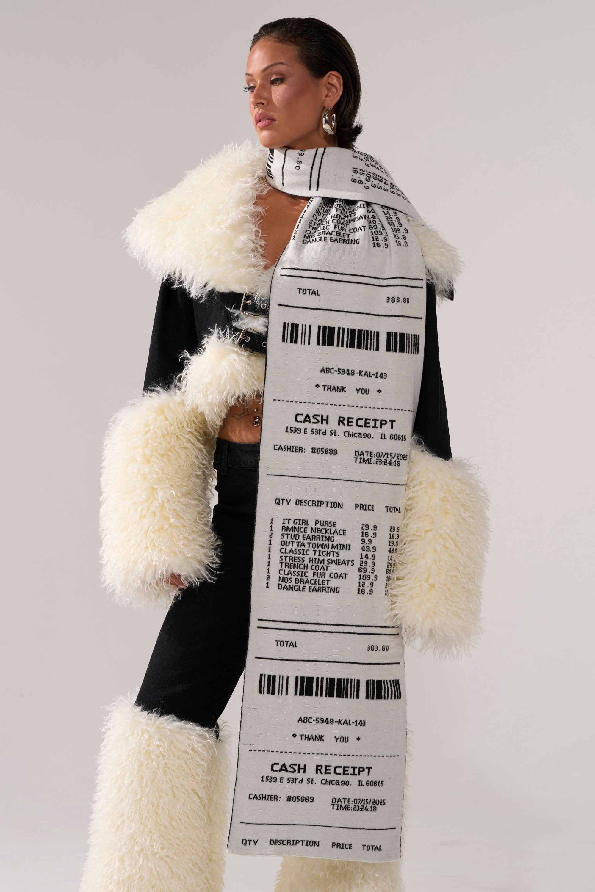 A woman in a black outfit with bold white faux fur cuffs and collar wears the CHECK THE RECEIPT SCARF—a playful wool-blend knit scarf styled as a printed shopping receipt—against a plain, light background. 3