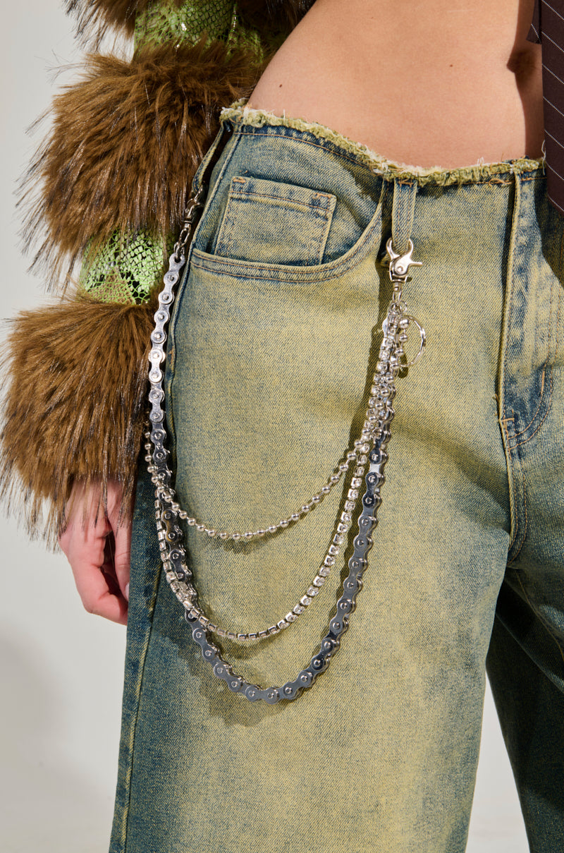 A person wearing the WHERES THE JUNK BAG PANT accessories with a purse chain draped along the pocket. The outfit includes a green and brown textured top with faux fur sleeves and a loosely hanging tie. These pants have a rugged waistline, adding an edgy vibe to the ensemble.
