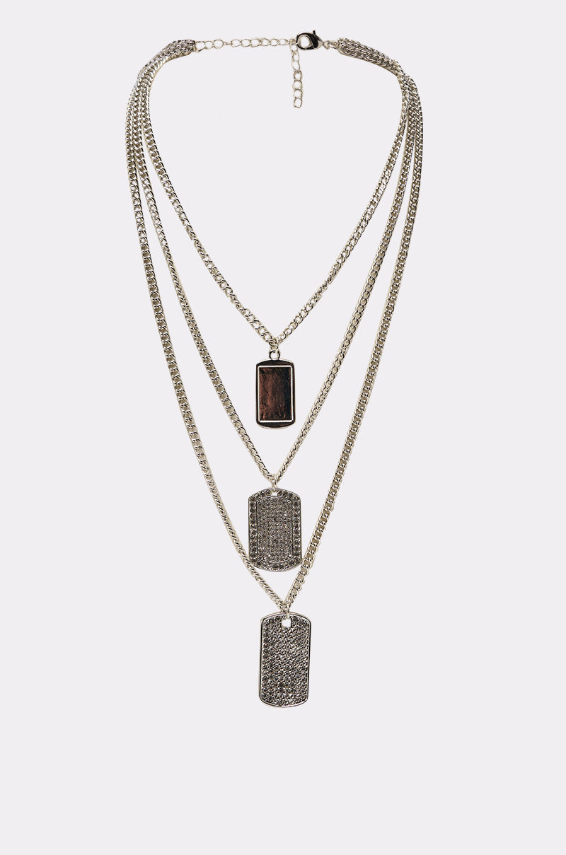 The DOG TAG BUT MAKE IT CUTE NECKLACE features three layered chains, each with a rectangular dog tag pendant: the top has a dark center, while the two lower tags display textured, silver-toned surfaces. 0