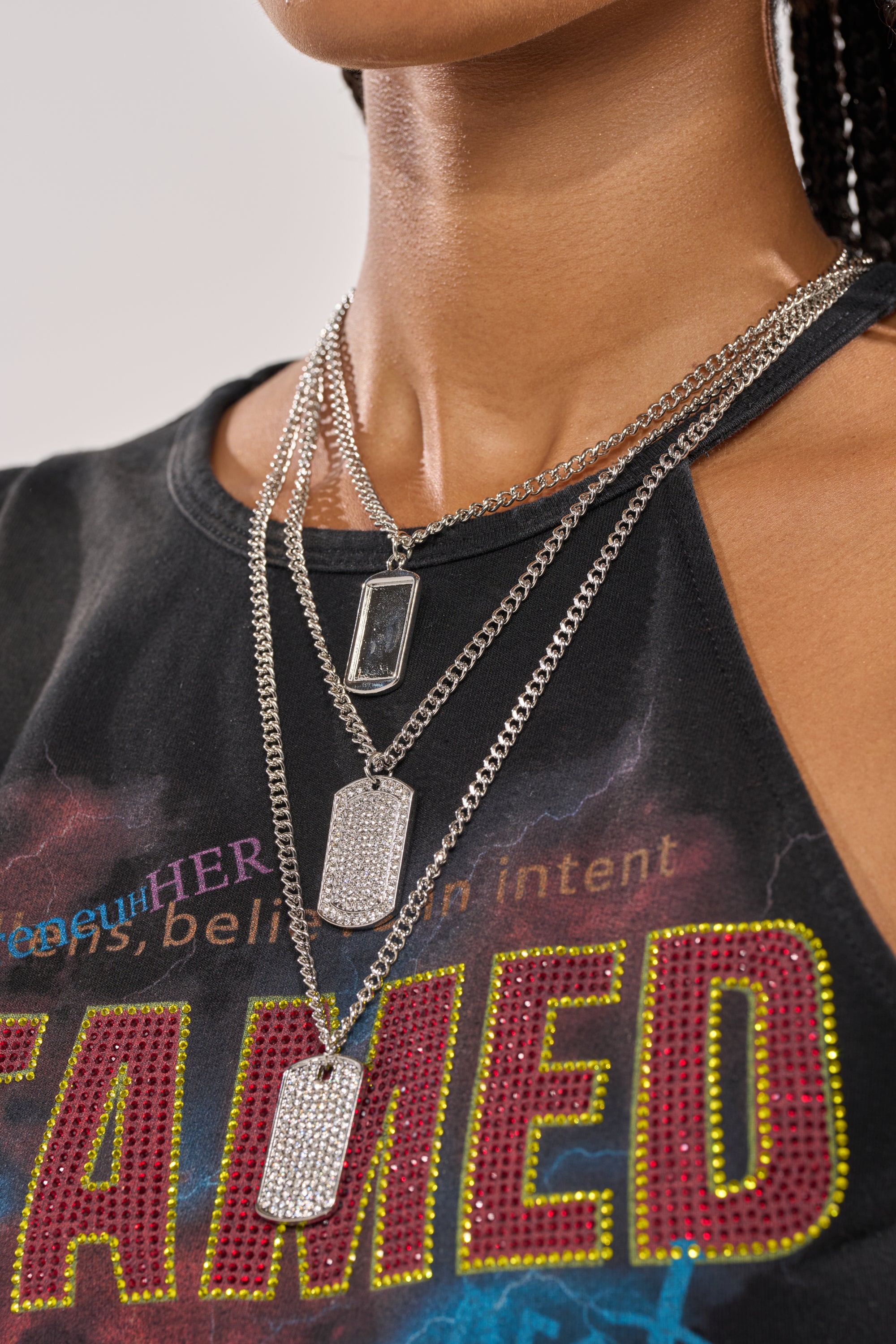 A person wears a black sleeveless shirt with colorful text and the DOG TAG BUT MAKE IT CUTE NECKLACE—three layered silver chains with rectangular and dog tag-style pendants—around their neck. 1