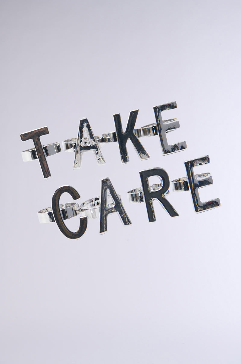 The faux silver "TAKE CARE" hairpins create a bold statement against a plain gray background, showcasing metallic letters in a modern and simple style.