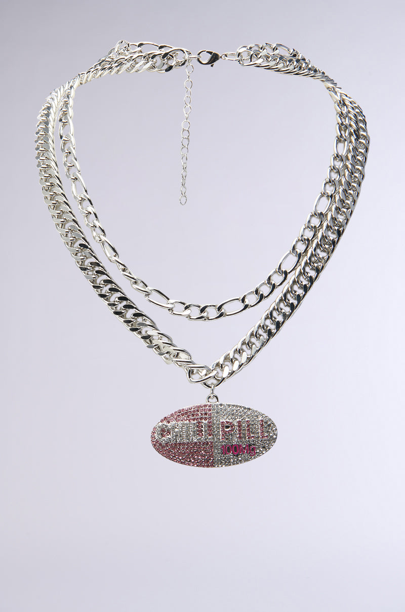 The TAKE A CHILL PILL NECKLACE is a statement piece, featuring a silver dual-layer chain and an oversized oval pendant reminiscent of a pill. Decorated with pink and clear rhinestones, it prominently showcases "CHILL PILL" in raised letters.