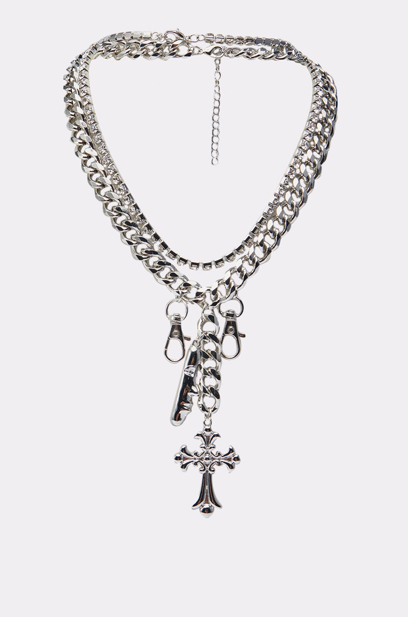 The ALL I SEE IS CHROME NECKLACE features three layered silver chrome chains—a Cuban chain, two lobster clasp charms, a whistle charm, and a decorative cross pendant at the center. 2