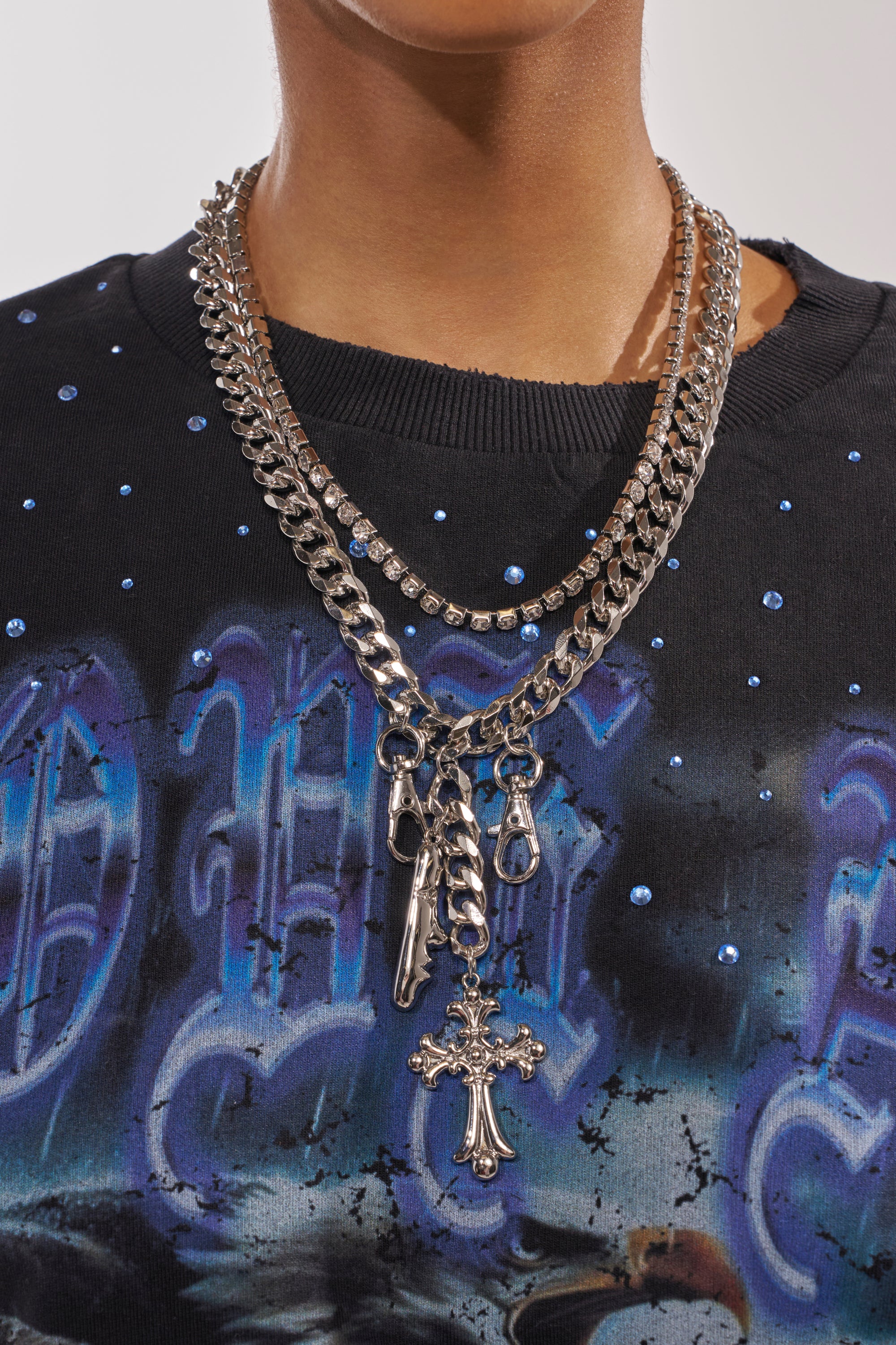 A person wearing the ALL I SEE IS CHROME NECKLACE—a layered silver chain with a cross pendant—over a black graphic shirt featuring blue designs and rhinestones. 1