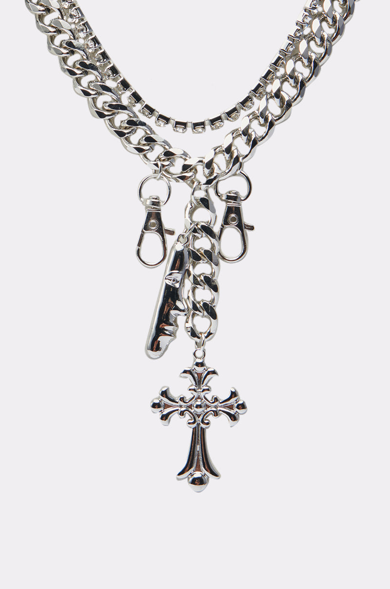 The ALL I SEE IS CHROME NECKLACE features three layered silver chains—a cross pendant, a Cuban chain, two lobster clasps, and a metal tag charm—all displayed on a plain white background. 3