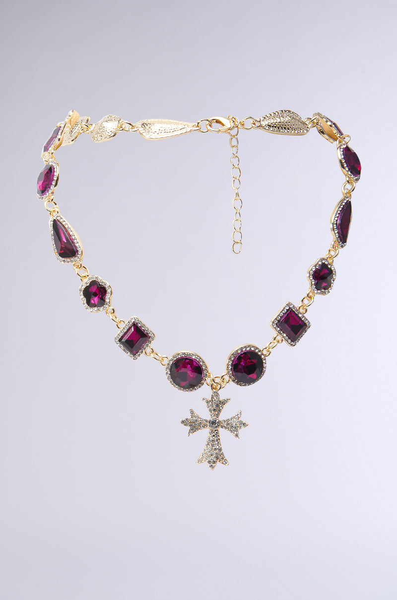 The INSPIRE ME CROSS NECKLACE by AKIRA Label features a silver cross pendant adorned with small diamonds and a rhinestone trim, showcasing an intricate design of variously shaped purple gemstones linked ornately. It has an adjustable chain clasp and is set against a soft gradient background.