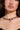 A woman is wearing the INSPIRE ME CROSS NECKLACE, showcasing purple gemstones and a small cross pendant. She has long dark hair and sports a black off-the-shoulder top with a subtle rhinestone trim. Her gold earrings complement the ensemble, while her lips are adorned with neutral lipstick.