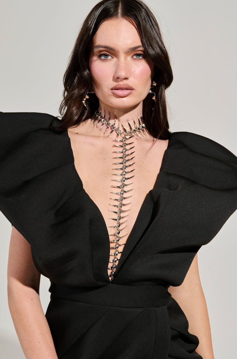A woman with long, dark hair poses in a striking black dress that has structured shoulder details and a deep V-neckline. She accessorizes with the ALL DOWN MY BACK STATEMENT NECKLACE, adorned with spiked charms, against a plain background to highlight this dramatic fashion piece.