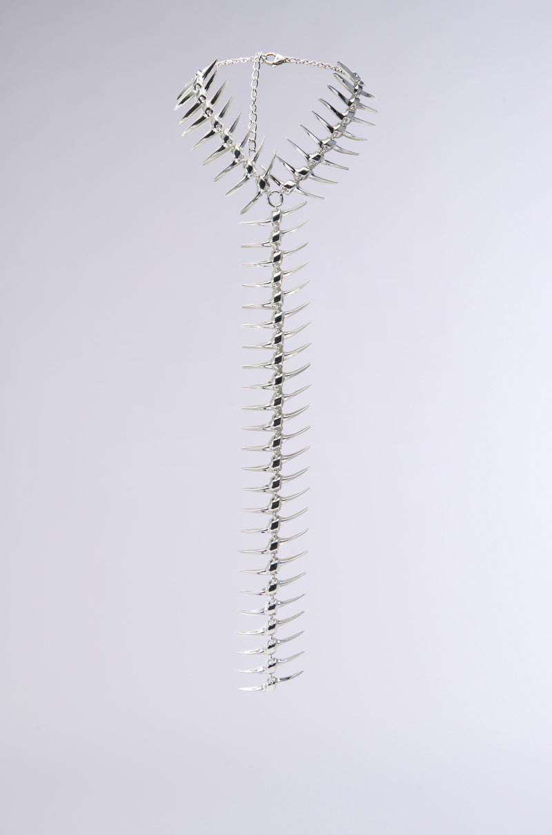 The ALL DOWN MY BACK STATEMENT NECKLACE is a striking silver piece with a long extension, adorned with numerous evenly spaced spiked charms for a bold and edgy appearance set against a neutral gray backdrop.