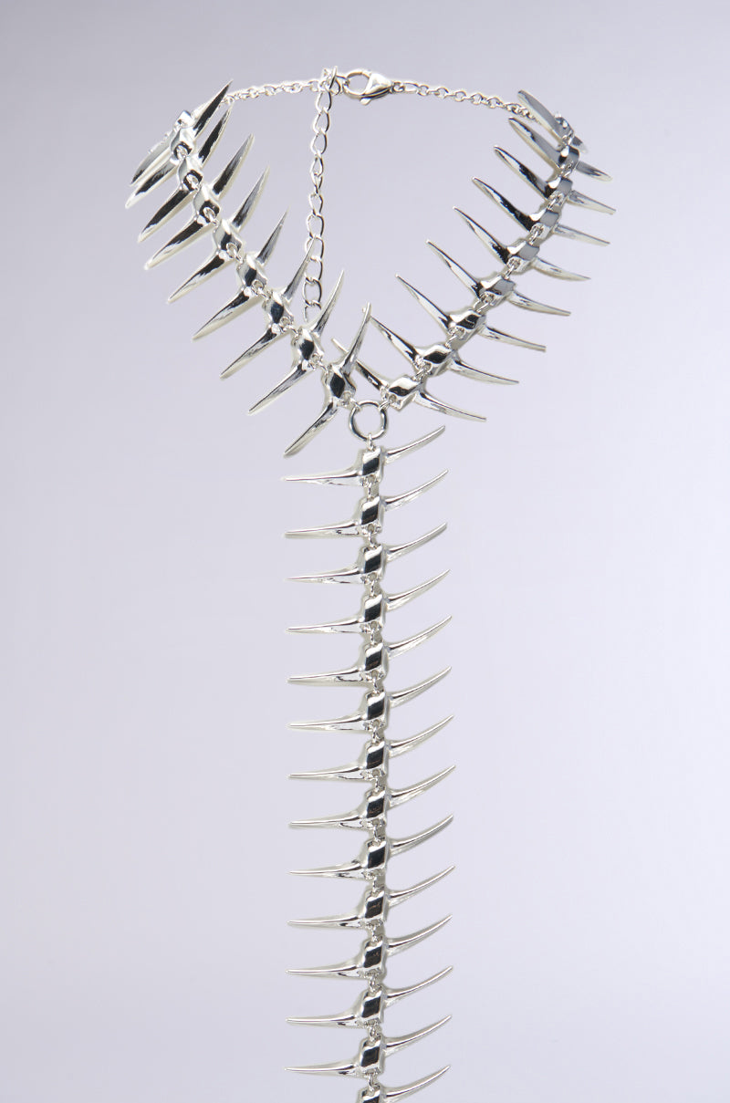 The ALL DOWN MY BACK STATEMENT NECKLACE features a striking, spiky design with numerous sharp, pointed metal components interconnected in a metallic chain. It is elegantly showcased on a light grey background.