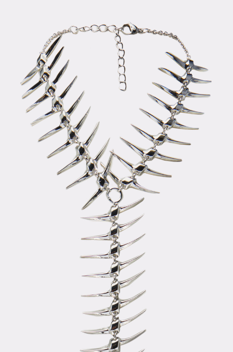 The ALL DOWN MY BACK STATEMENT NECKLACE features a metallic silver, V-shaped fish skeleton design with pointed segments and spiked charms on a chain link, finished with an adjustable clasp.