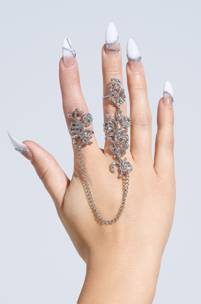 A hand adorned with the GALA BOUND RING SET, featuring intricate floral patterns and a chain linking two fingers, flaunts manicured nails showcasing a white and clear marble design on smooth, light skin.