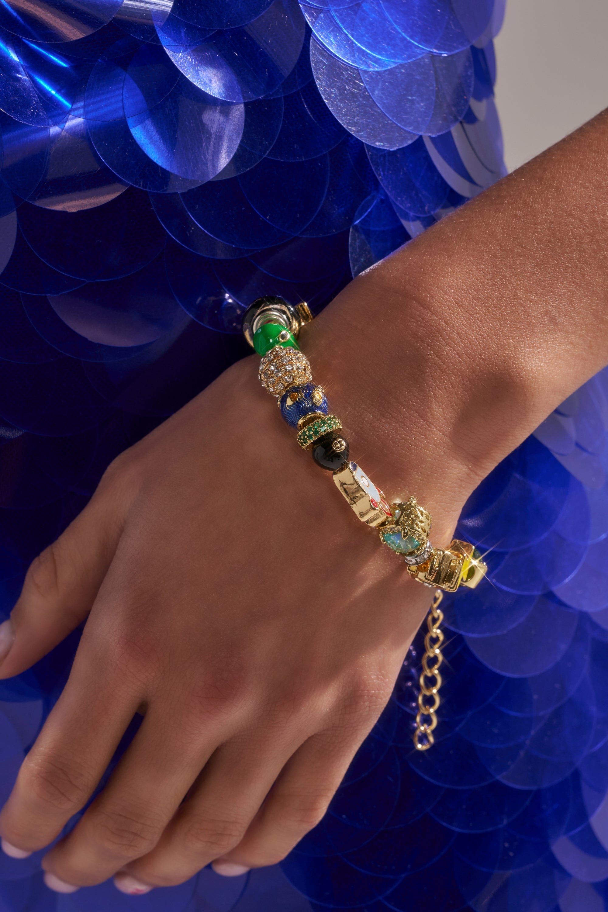 A hand wears the SPACED OUT CHARM BRACELET, a celestial jewelry-inspired gold-tone piece with colorful charms and crystals, resting against a dress decorated with large, shiny blue sequins. 1