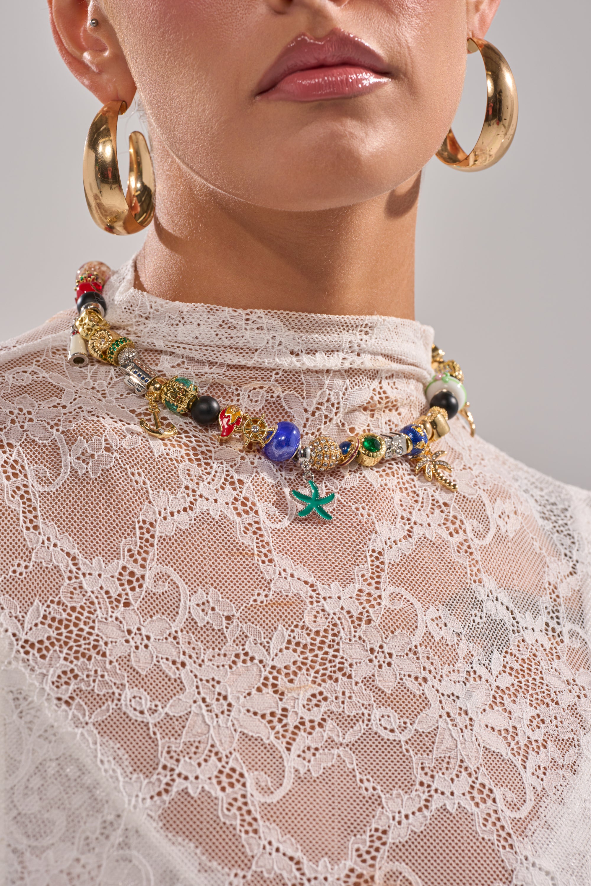 A woman wears large gold hoop earrings and the THIS AND THAT NECKLACE, a playful statement piece with colorful beads, over a high-neck white lace top. Only her lower face, neck, and part of her upper body are visible.