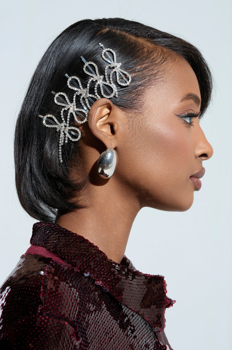 A side profile of a woman showcases her short, sleek black hair embellished with the intricate COQUETTE DREAMIN RHINESTONE CLIPS. She is dressed in a maroon sequined top and accessorizes with large silver earrings against a plain, light-colored background.