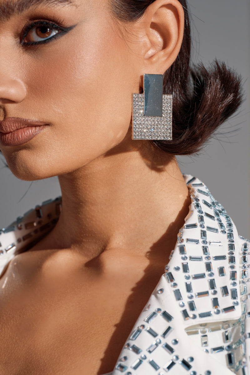 A close-up of a woman wearing the JESS STATEMENT EARRING—bold, geometric silver earrings—paired with a white jacket featuring silver studs; her hair is slicked back and she sports dramatic black winged eyeliner.