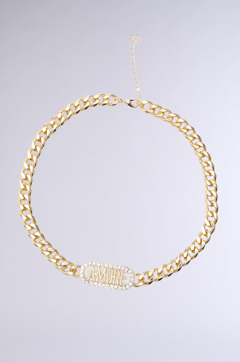 The LOVE IS IN THE AIR NECKLACE IN GOLD features a chunky curb chain with a rhinestone centerpiece spelling "AMORE," surrounded by small gemstones. It includes a small adjustable clasp and chain extender, resting on a neutral gray background.
