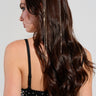 A woman with long, wavy brown hair is facing sideways. She is wearing the ON MY MIND BLING HEADBAND, which features a rhinestone fringe and strands of gold accessories shimmering through her hair, along with a black sleeveless top adorned with silver studs, creating an enchanting effect.