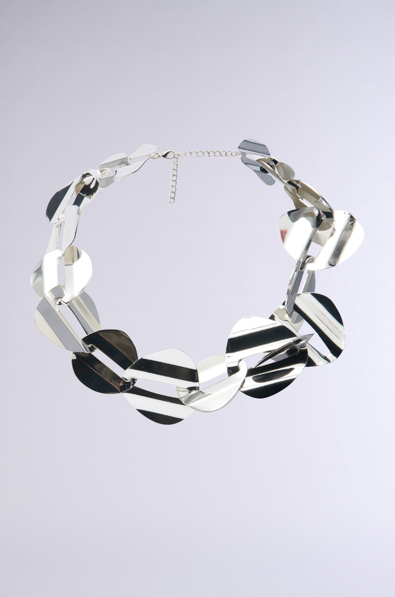 The IYKYK STATEMENT NECKLACE showcases a striking choker design with interlocking geometric links, creating a sleek and modern reflective look. It features an adjustable chain link strap at the back and is displayed against a simple, light backdrop in the photograph.