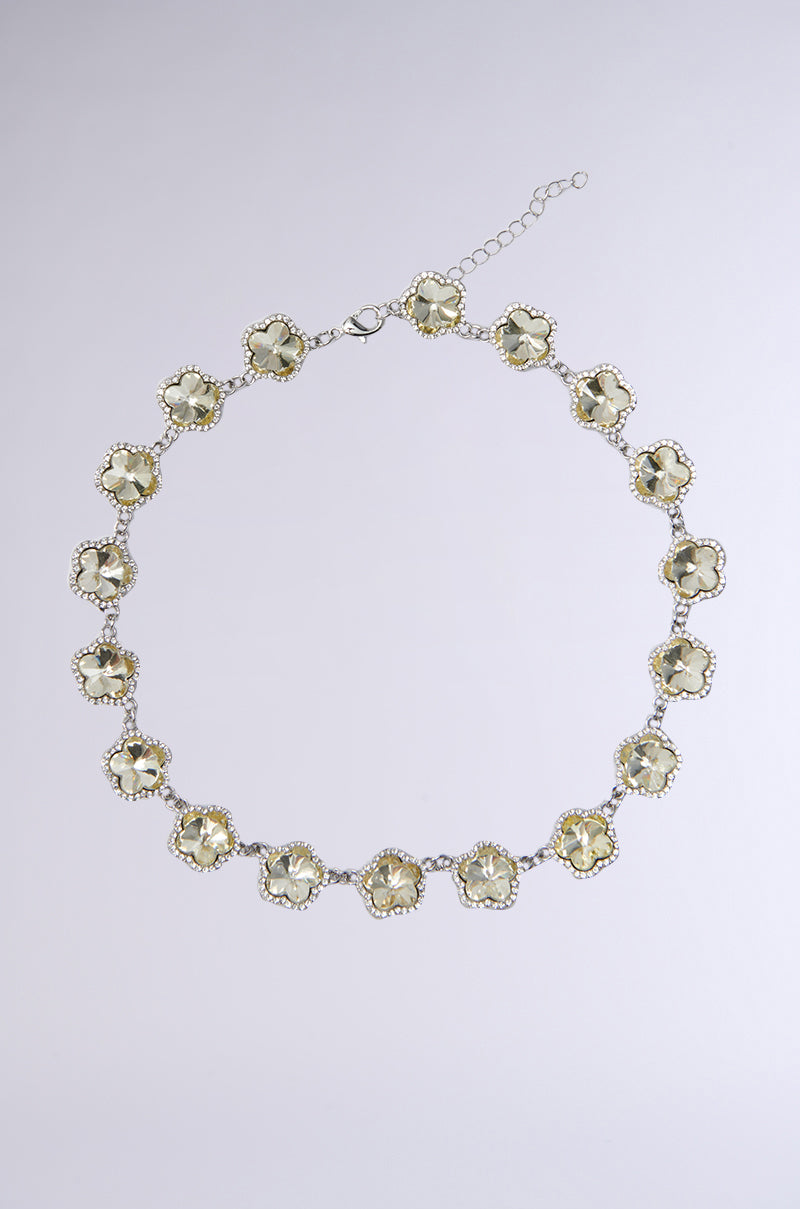 The QUEZ COULD NEVER NECKLACE showcases circular, flower-like patterns with light yellow gemstones and a pave rhinestone trim. Set in silver-toned metal, it has an adjustable chain clasp. Its elegance is highlighted against a plain gray background.
