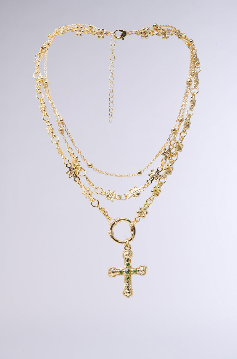 The PRAYING FOR SPRING LAYERED NECKLACE, a faux gold piece with intricate designs, is displayed on a light grey background. It features multiple chains and an elegant standout cross pendant at the center.