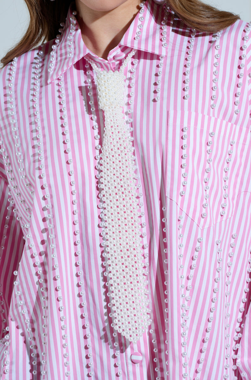 The PEARLY TIE shirt features pink and white stripes with faux pearl beads and a textured necktie silhouette, showcasing an elegant design against a plain background.