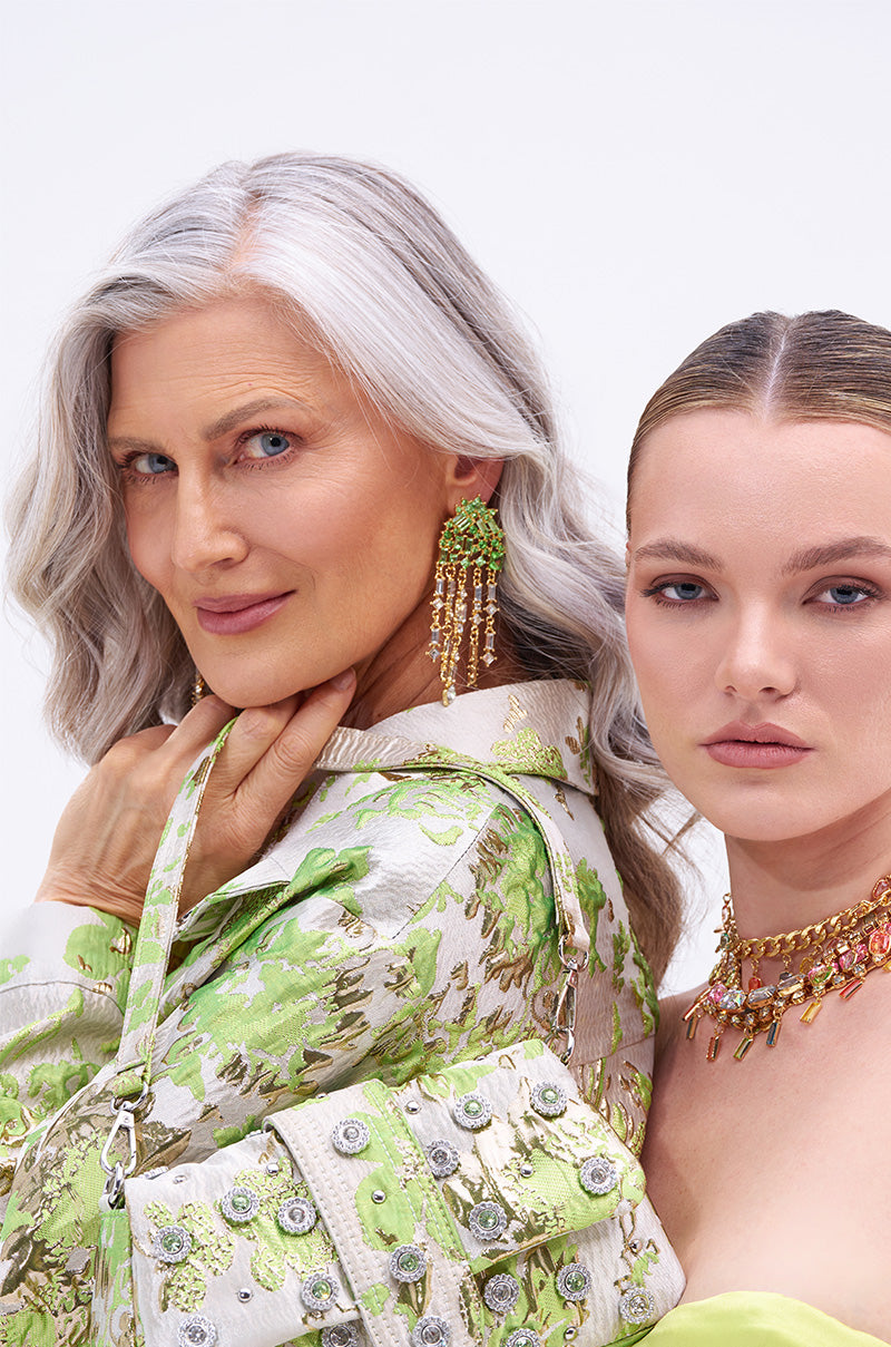 Two women pose before a light background. The older woman, in green floral clothing, wears the BLING ME HOME EARRING with a cascading dangle look. The younger woman complements her pastel outfit with a gold necklace. 1