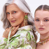 Two women pose before a light background. The older woman, in green floral clothing, wears the BLING ME HOME EARRING with a cascading dangle look. The younger woman complements her pastel outfit with a gold necklace.