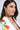 A woman with long brown hair models the BLING ME HOME EARRING, a cascading dangle design with bold gold and emerald green stones, styled with a white floral blazer as she gazes to the side in bright light.