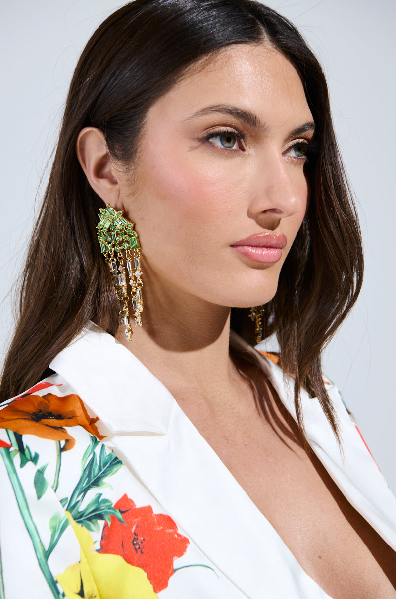 A woman with long brown hair models the BLING ME HOME EARRING, a cascading dangle design with bold gold and emerald green stones, styled with a white floral blazer as she gazes to the side in bright light. 2