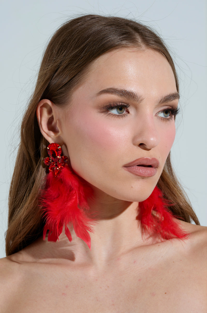 A woman with long brown hair wears striking SEE RED FEATHER EARRINGs and subtle makeup. Her expression is neutral, drawing attention to her earrings. The background is plain and light-colored.