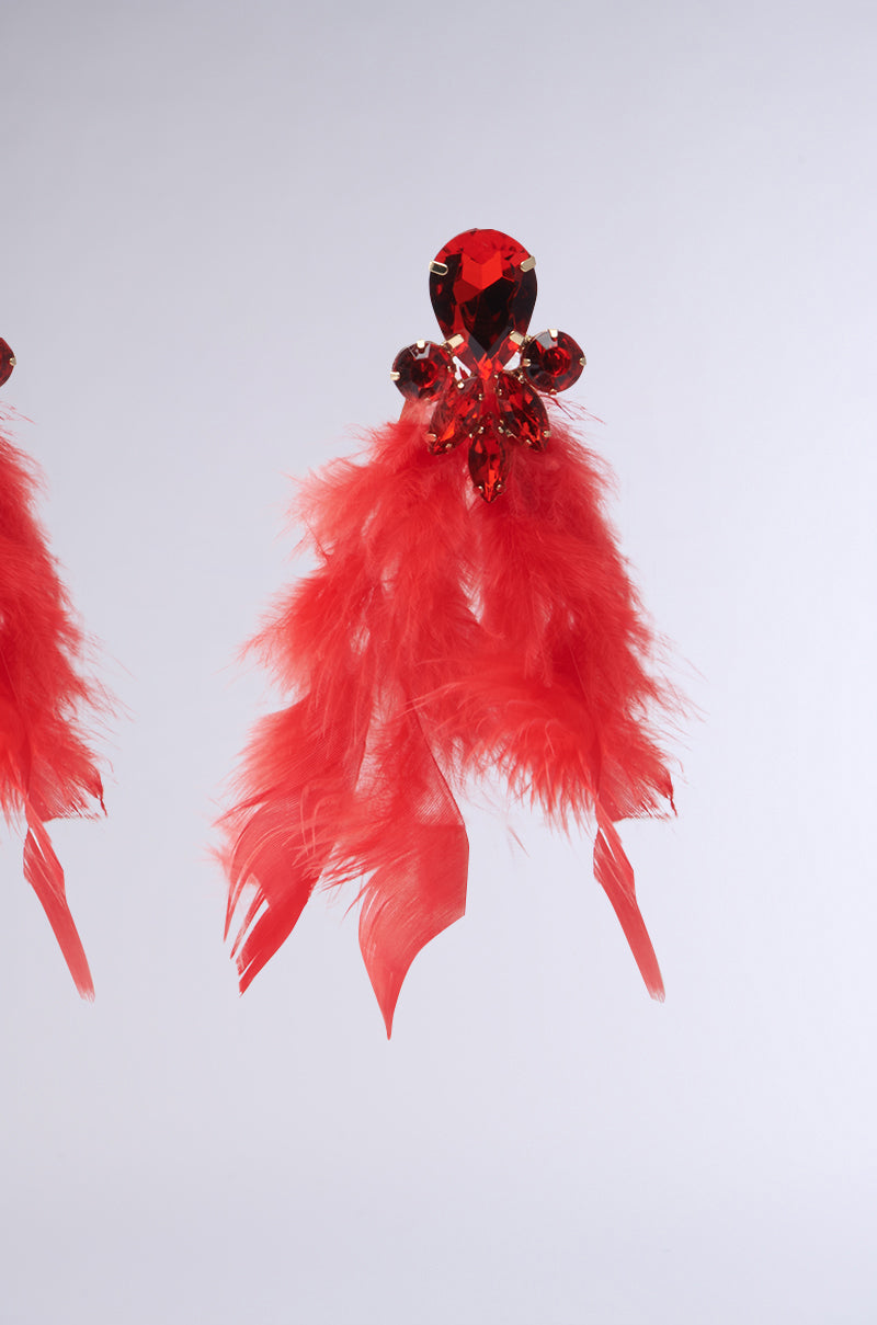 Close-up of the SEE RED FEATHER EARRING from AKIRA Label, showcasing a ruby-inspired teardrop gemstone encircled by smaller stones and adorned with striking red feathers cascading down. Set against a plain, light gray background, highlighting its intricate details and vibrant colors.