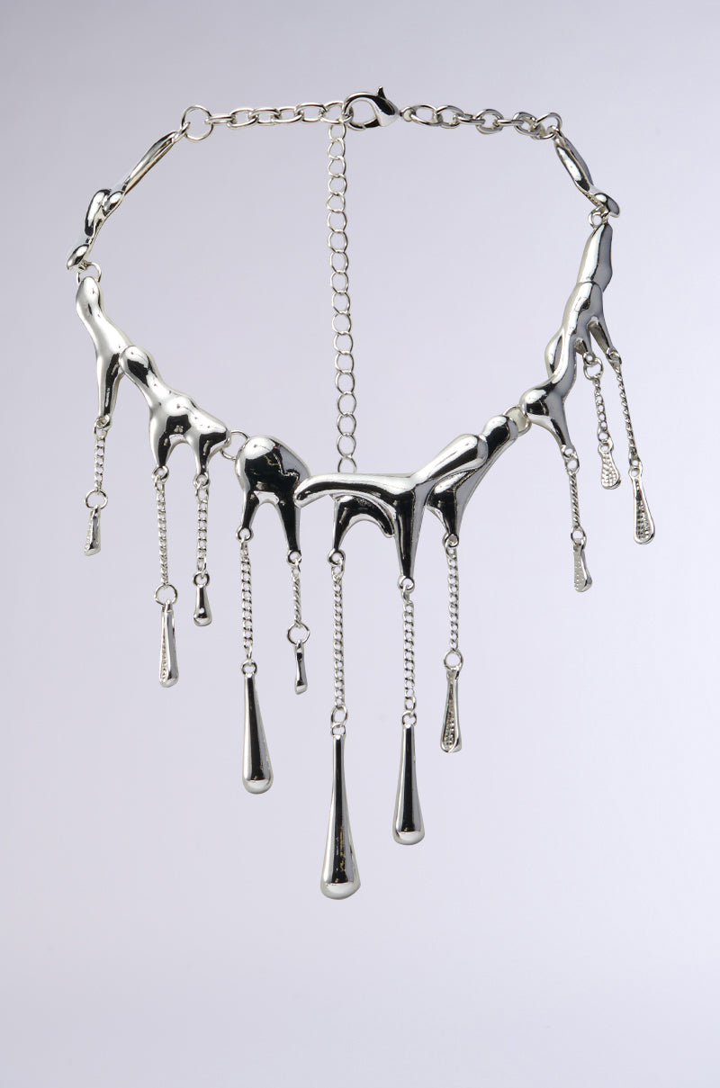 The BRICK WALL WATERFALL NECKLACE is a shiny silver piece with a molten metal design, featuring teardrop embellishments hanging from cascading chain links of varying lengths, set against a plain, neutral background.