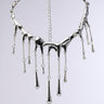 The BRICK WALL WATERFALL NECKLACE is a shiny silver piece with a molten metal design, featuring teardrop embellishments hanging from cascading chain links of varying lengths, set against a plain, neutral background.