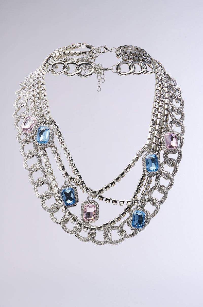 The PASTELS PLS NECKLACE SET, a chunky chain multi-layered silver necklace with large rectangular gemstones in pastel blue and pink, is displayed against a plain gray background.