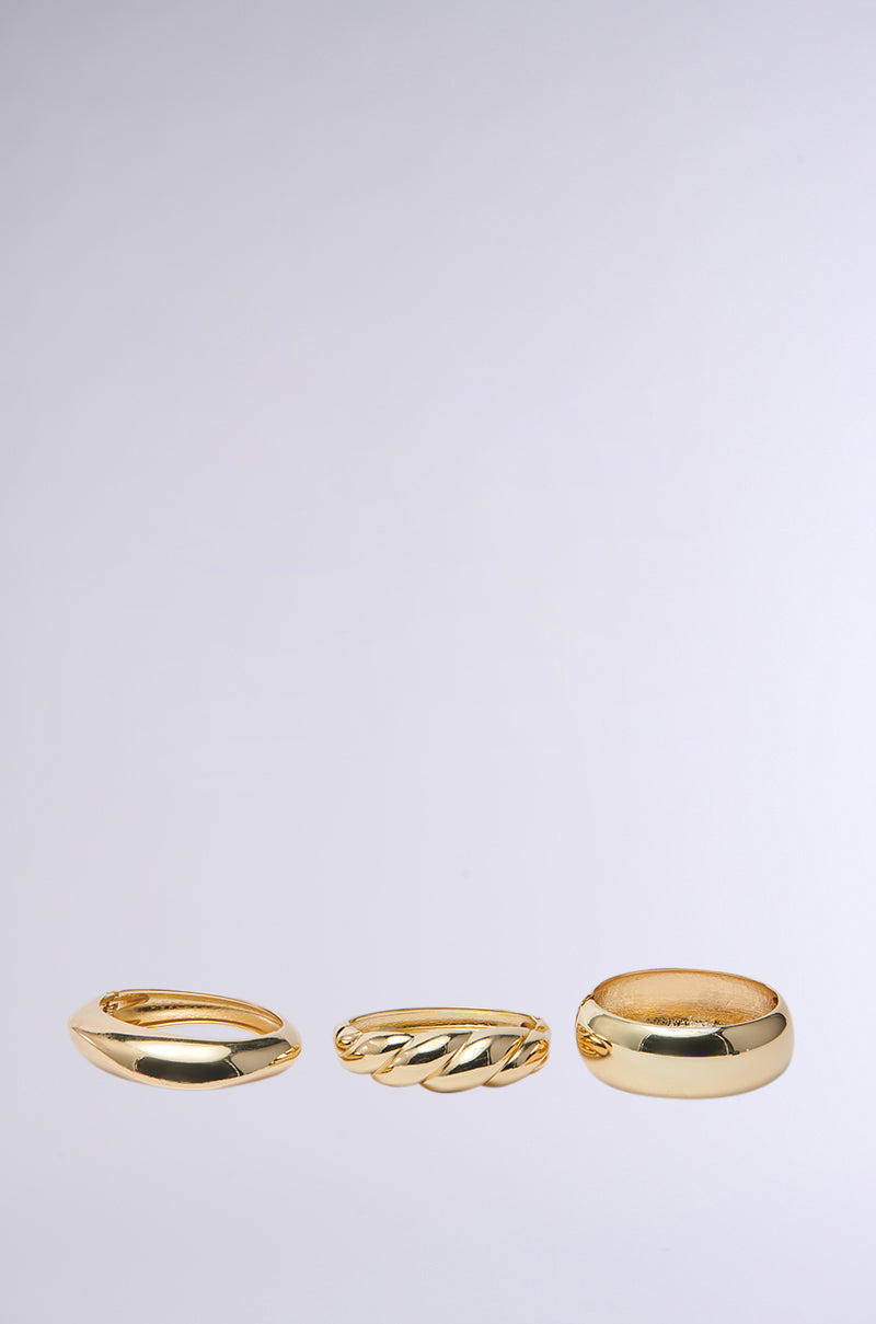 The BITE ME GOLD BRACELET SET, featuring three faux gold rings, is showcased on a light gray background. The set includes a plain, slightly curved ring, a twisted pattern ring, and a smooth wide band resembling chunky bangles. 1