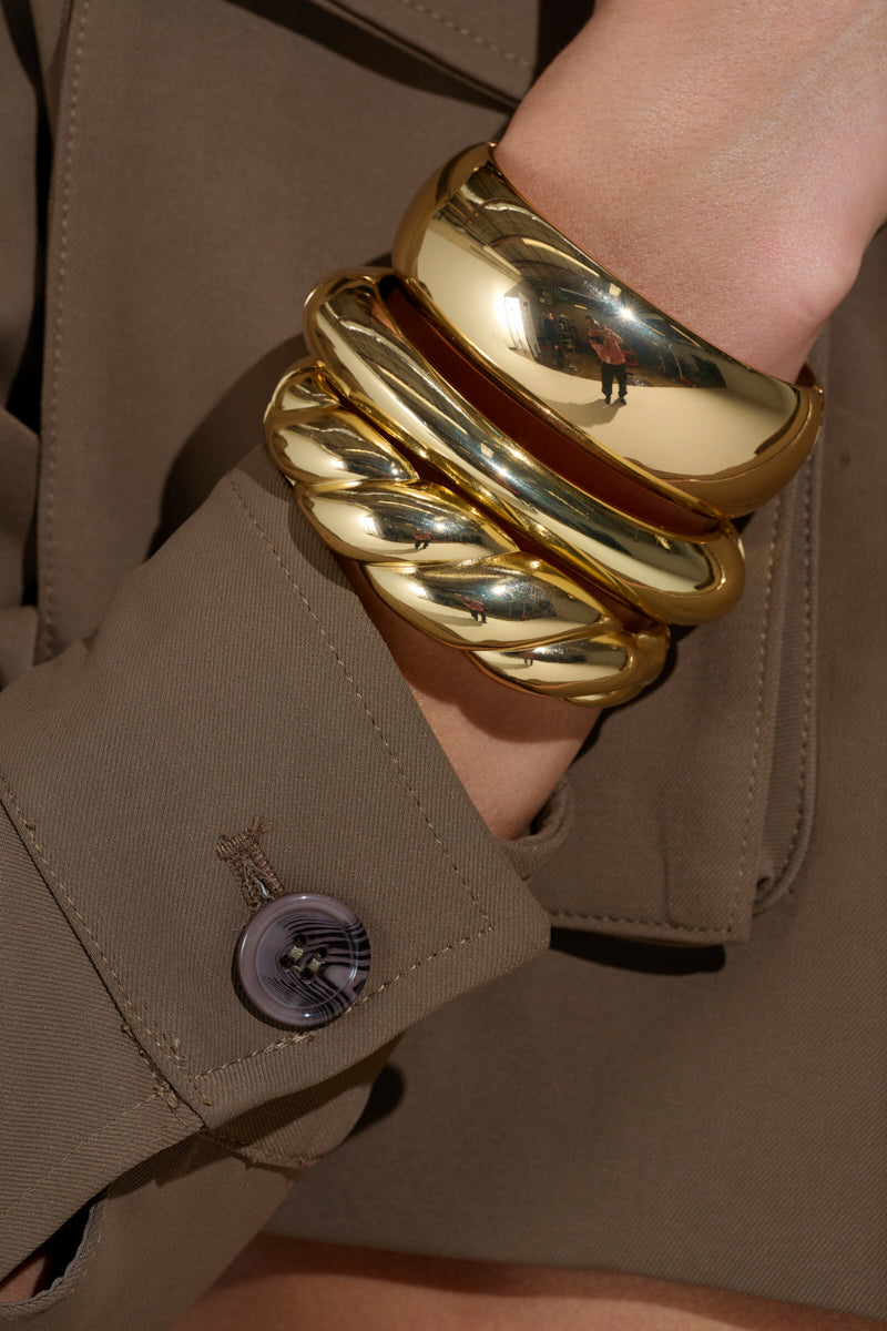 A close-up shows a wrist adorned with the BITE ME GOLD BRACELET SET. The chunky faux gold bangles, featuring two smooth designs and one twisted texture, are elegantly paired with a brown jacket, highlighting their style next to a visible cuff button. 0