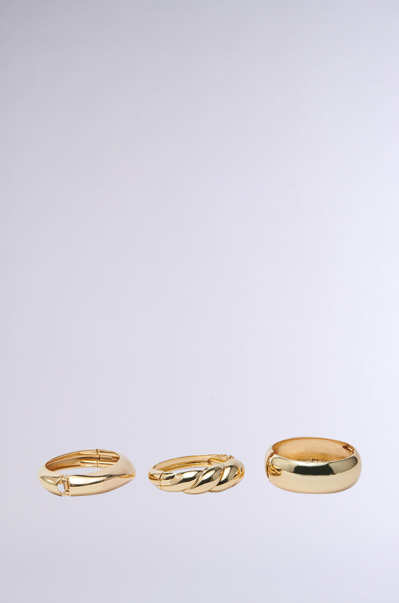 The BITE ME GOLD BRACELET SET showcases three faux gold rings. The left ring features a slim, smooth design; the middle one has a braided pattern; and the right ring is wider with a polished finish, reminiscent of chunky bangles, all set against a plain background. 2