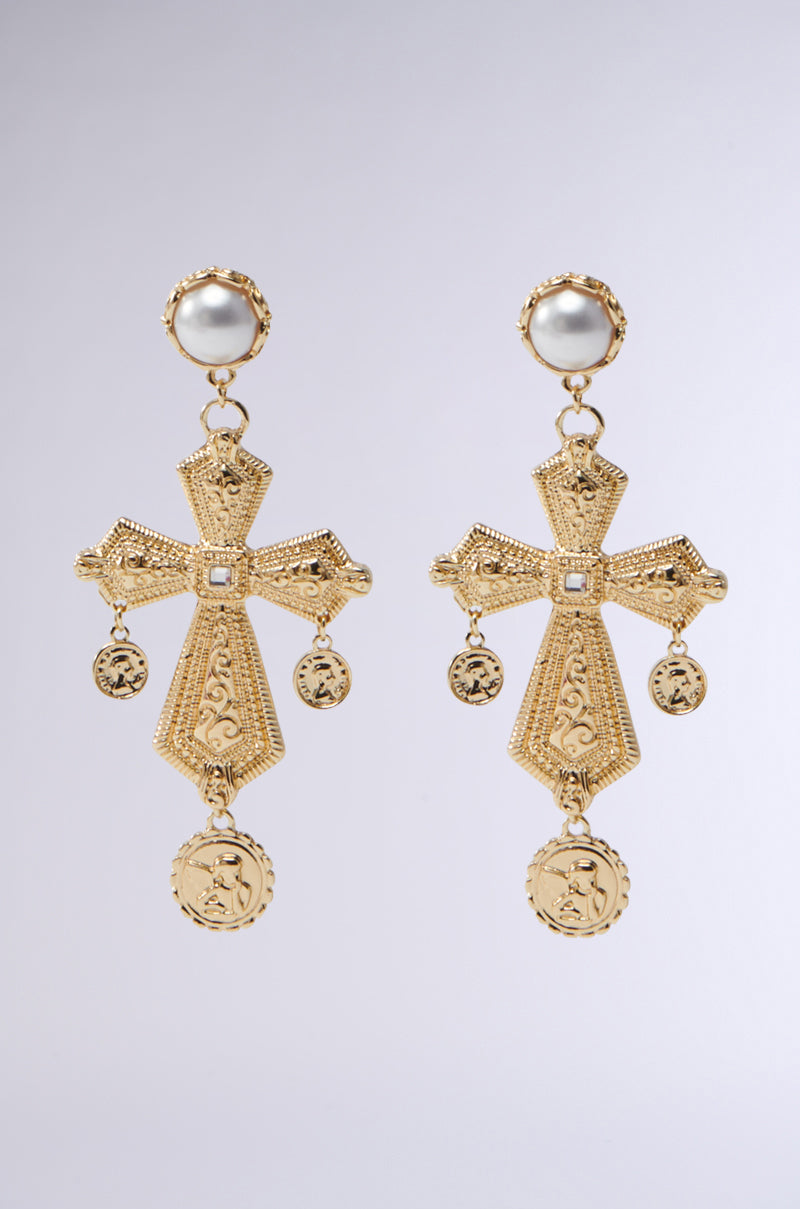 The PRAY FOR U EARRING by the AKIRA Label features gold cross-shaped designs with intricate patterns and elegant pearl posts. Each earring showcases dangling gold coins with embossed designs, set against a simple gray gradient background.