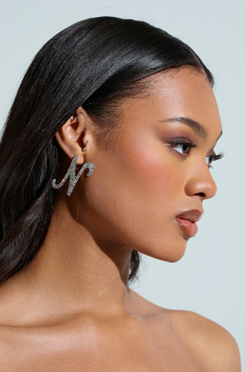 A person with long, dark hair is shown in profile against a light background, wearing the striking NO THX EARRING adorned with rhinestone "M" embellishments. Their makeup includes subtle eyeshadow and blush. 1