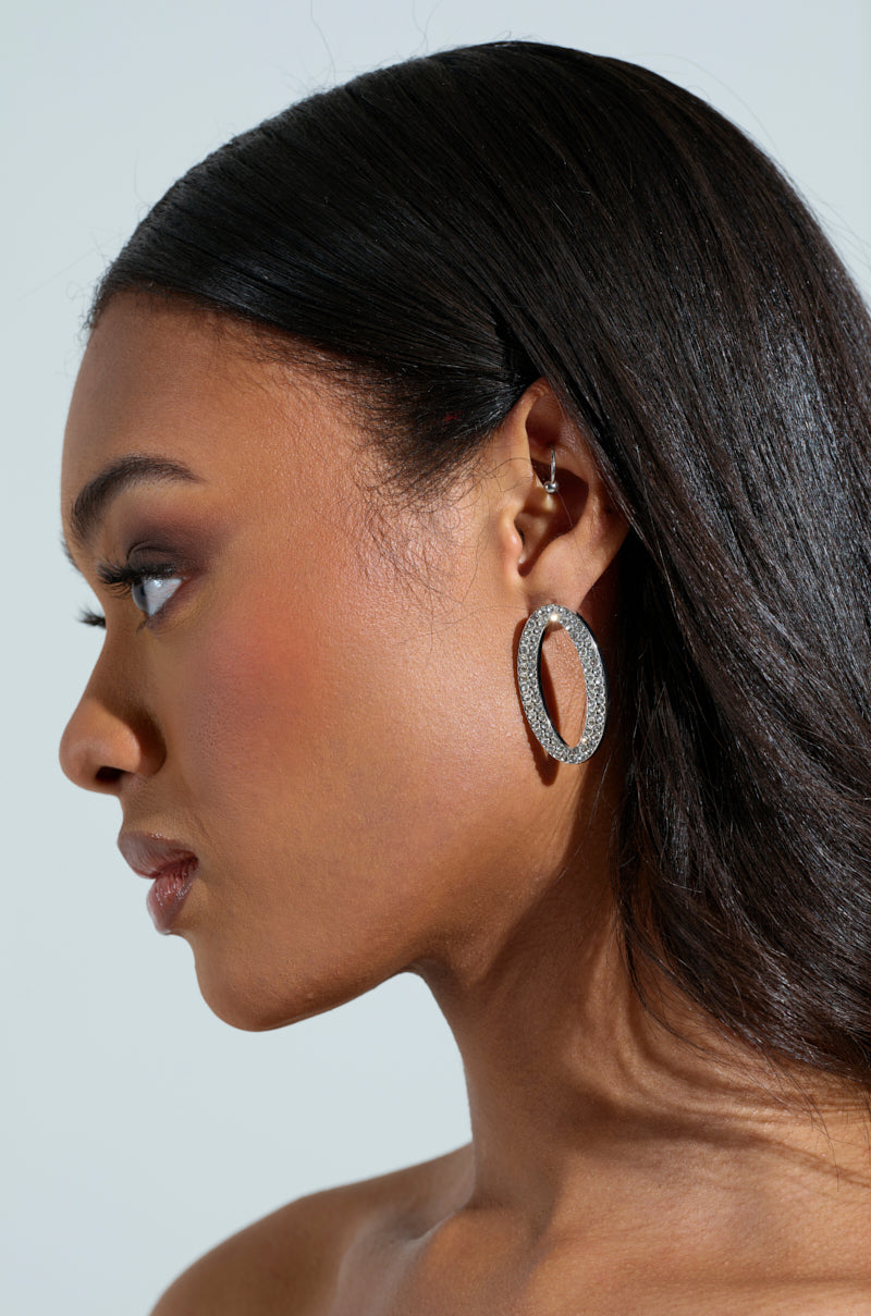 Side profile of a person with long dark hair, wearing the NO THX EARRING, a sparkly faux silver metallic oval-shaped hoop with rhinestones and a small ear cuff on the helix. The plain background highlights the jewelry and the person's features. 2